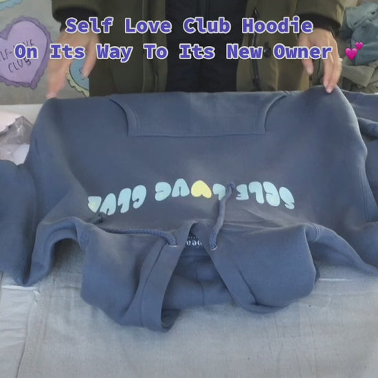A person folding an ocean colored "SELF LOVE CLUB" hoodie and packaging the hoodie in a shopping bag.
