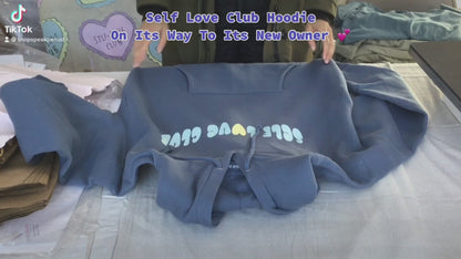 A person folding an ocean colored "SELF LOVE CLUB" hoodie and packaging the hoodie in a shopping bag.