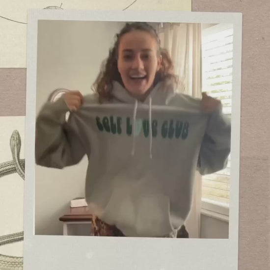 A girl in a sage colored "SELF LOVE CLUB" hoodie smiling, tilting her head, and showing off the design of the hoodie. 