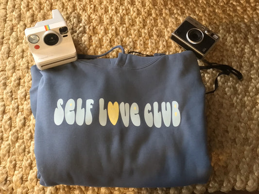 A folded ocean colored "SELF LOVE CLUB" hoodie next to polaroid cameras.
